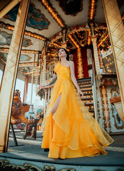 Young beautiful woman in a bright yellow evening dress posing against the backdrop of an amusement park and carousel — Stock Photo, Image