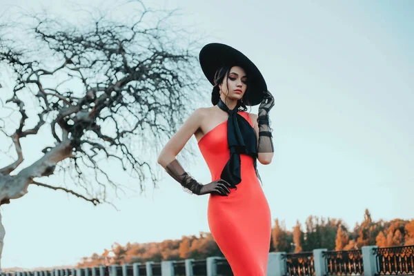 Elegante Retro-Lady in einem langen roten Abendkleid. Junge, stylische Trendfrau mit schwarzem Hut mit breiter Krempe und hohen Tüll-Handschuhen. Verbeugung am Hals. Erwachsenes Modelmodel steht auf der Straße — Stockfoto