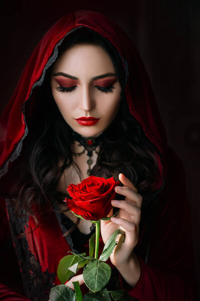 Mystical beautiful woman in a gothic costume of a medieval vampire in a hood. An adult girl holds a rose flower in her hands. Beautiful face red makeup, attractive lips. Halloween party image