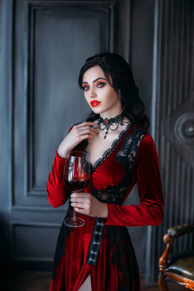 Hermosa joven mujer sexy vampiro en el castillo medieval oscuro. Vestido de noche gótico rojo. Pelo ondulado negro. Cuarto vintage telón de fondo. Una chica sosteniendo una copa de vino sangriento en sus manos. Imagen reina de la noche —  Fotos de Stock