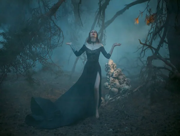 Розмитий силует містичної язичницької жінки в тумані. Відьма кидає закляття, молиться в лісі руками, піднятими до неба на тлі темного дерева і людських черепів — стокове фото