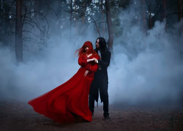 Silhueta turva de um casal gótico dançando no nevoeiro. Um homem vampiro de casaco preto com cabelo comprido seduz uma mulher com um longo vestido medieval vermelho. O tecido e o cabelo estão voando no vento — Fotografia de Stock