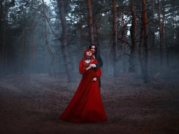 Casal gótico parado no nevoeiro. Um homem vampiro de casaco preto com cabelo comprido abraça uma mulher com um longo vestido medieval vermelho. Foto de arte de fantasia. Fundo floresta escura profunda, árvores, nevoeiro azul. — Fotografia de Stock