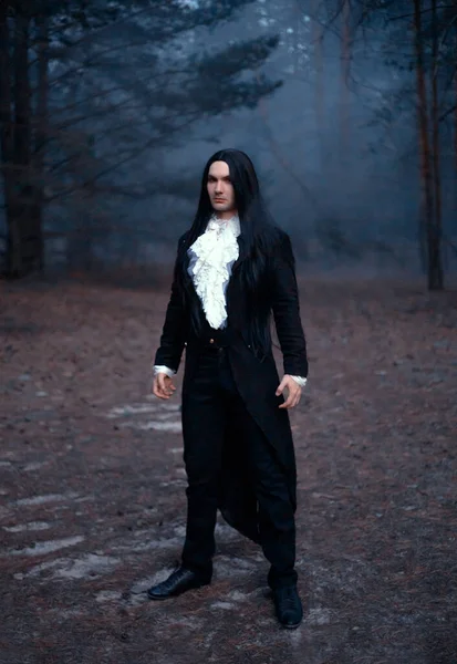 Egy férfi Drakula gróf képében fekete kabátban. Gótikus jelmez fehér vintage frillel. Hosszú haj, középkori frizura. Természeti háttér, misztikus sötét erdő. Dühös arc sápadt bőr vörös ajkak — Stock Fotó