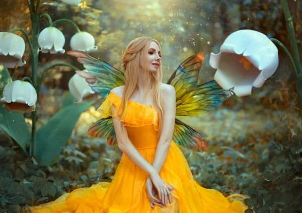 Portrait de femme heureuse fantaisie blonde fée de la forêt. Modèle de mode dans une robe jaune vif avec des ailes de papillon est assis posant dans la nature. De grandes fleurs décor décor lys blancs. Éclat magique léger. — Photo