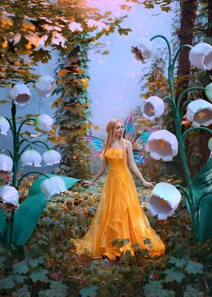 Une femme fantasmée fée de la forêt. Modèle de mode en robe jaune avec des ailes de papillon se promène dans la nature d'automne. De grandes fleurs décor décor lys blanc de la vallée. Feuilles orange de brouillard mystique bleu arbre. — Photo