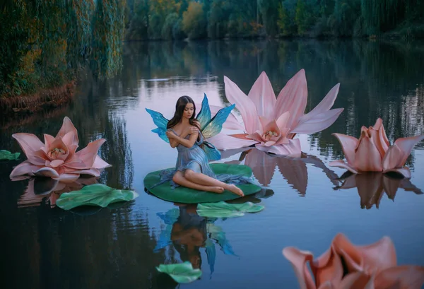 Una mujer hermosa un pequeño hada fabulosa con alas de mariposa se sienta en la hoja de lirio de agua verde. Escenario de fantasía de enormes flores rosadas en el lago, árboles verdes. Río ninfa inocente chica en un vestido azul —  Fotos de Stock