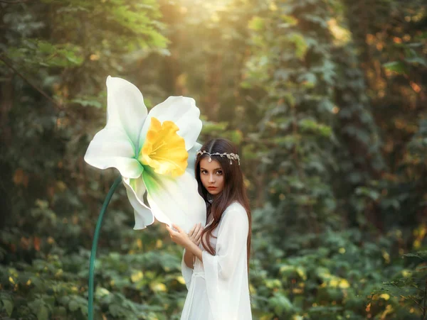 Porträt eines schönen Mädchens Elfe mit langen Haaren steht in einem Fantasiewald und umarmt eine riesige Narzissenblume, grüne Bäume. Eine Frau in weißem Vintage-Kleid und silbernem Diadem. Unschuldiges attraktives Gesicht — Stockfoto