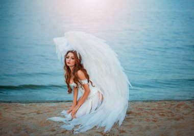 Genç ve güzel bir kadın, düşmüş üzgün bir melek, deniz sahilinde oturuyor. Yaratıcı seksi kostüm, büyük yapay kuş kanatları ve beyaz vintage elbise. Pişman suratlı yetişkin bir kız. Resim fotoğrafı.