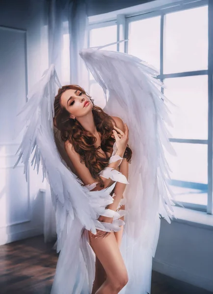 Jovem bela mulher anjo com enormes asas brancas fica na sala perto da janela. — Fotografia de Stock
