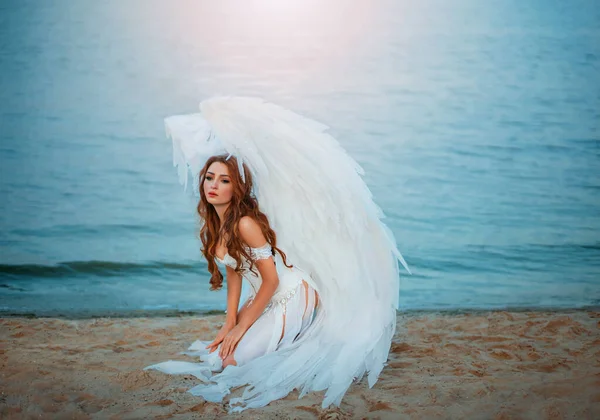 아름다운 여인이 죽어 가는 슬픈 천사가 해변에 앉아 있다. 창조적 인 섹시 한 의상, 거대 한 인공 새 날개, 그리고 흰색 빈티지 드레스. 회개의 슬픈 얼굴을 한 성인 소녀. Artwork photo — 스톡 사진