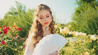 Portre klasik bayan ortaçağ prensesi elinde yelpaze sallıyor serinliğin tadını çıkarıyor taze hava yeşil yaz rüzgarı doğa gülleri. Genç kadın, sarışın, eski tarz beyaz takı seti.