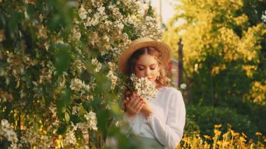 Sanat portresi modeli yetişkin mutlu kız eli yeşil yapraklara dokunuyor beyaz çiçek ağacı. Retro Bayan Güzellik, yaz bahçesi aromasının tadını çıkarın. Genç bayan, uzun kızıl saçlı, eski moda şapka 4K.