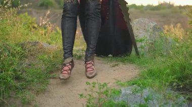 Fantezi kadın savaşçı yürüyor. Dişi bacakları patika boyunca yaklaşır. Seksi kız elinde metal altın bir kılıç tutuyor. Yaz doğası yeşil çimenler, yumuşak gün batımı. yaratıcı tasarım hight ayakkabı 4k