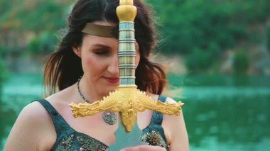 Savaşçı fantezi kadın savaş öncesi dua eder. Kılıcı ellerinde tutar. Güçlü yüz, yakın plan rüzgar saçlarını dalgalandırır. Kız İskandinav tarzı Valkyrie tanrıçası. Deri kostüm. Yeşil doğa nehri. video 4k