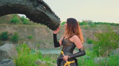  Kılıç tutan fantezi kadın savaşçı ejderha kafasına dokunuyor. Yeşil çimen dağları yaz doğası. Portre Savaşçısı Kadın Kraliçe seksi kostüm yaratıcı kıyafet deri zırh bronz zırh zırhı