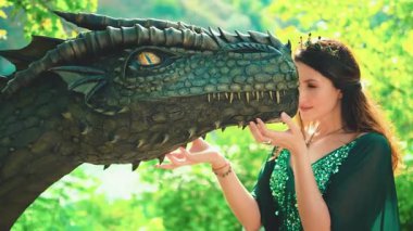 Fantezi kraliçesi evcil ejderhaya annesi gibi sarılıp dokunur. Sihirli ilahi güneş ışığı yaz ağaçları yeşil yapraklar. Elf elbisesi. Prenses tanrıça seksi manken kız. Efsane hayvan. 4k