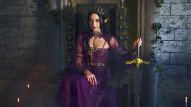 Sanat fantezi perisi vampir kadın şeytani elf kraliçesi elinde kılıç, koyu mor elbiseyle tahtta oturuyor. Seksi cadı elf kızı, sivri kulaklı. Gotik savaşçı prenses, altın taç. Eski tarz gece odası.