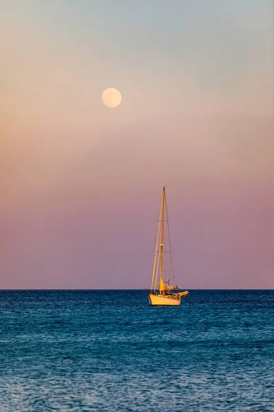 Full moon rising over the water with a small sailing boat in the foreground. Sailing boat with raising moon at sunset. Moon rising over the sea and yacht floating on the water surface. Sardinia, Italy