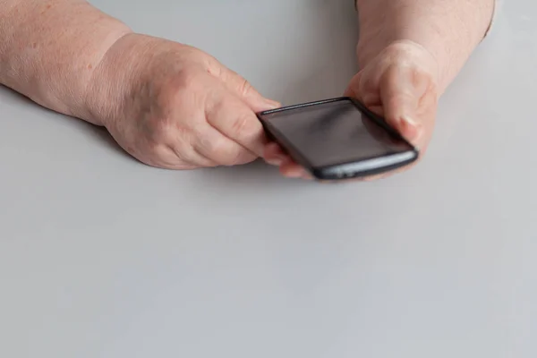 Black touchscreen phone in elderly hands on a gray background.