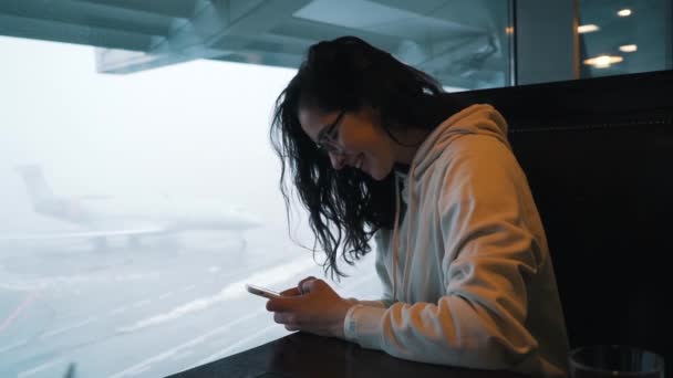 Menina com um smartphone senta-se no terminal do aeroporto — Vídeo de Stock