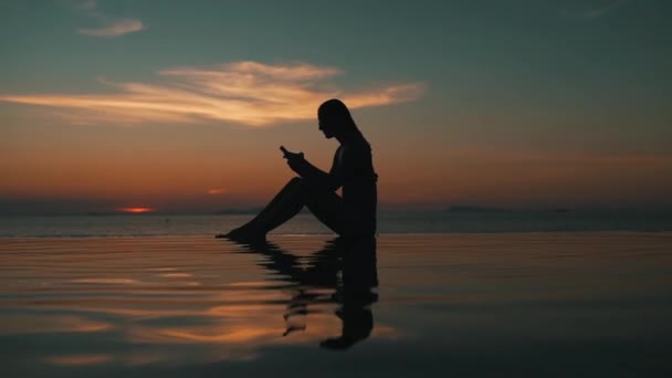Flickan använder en smartphone vid solnedgången vid havet — Stockvideo