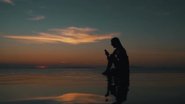 Chica utiliza un teléfono inteligente al atardecer por el océano — Vídeos de Stock