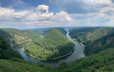 Kısmen bulutlu gökyüzünün altında kıvrılan nehri çevreleyen yemyeşil tepelerin panoramik manzarası