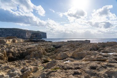 Parlak gökyüzü altında kayalık kıyı manzarası bulutlar ve güneş ışığı suya yansıyor