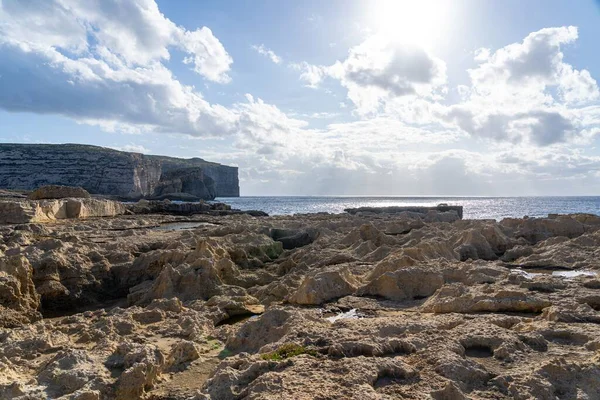 Parlak gökyüzü altında kayalık kıyı manzarası bulutlar ve güneş ışığı suya yansıyor