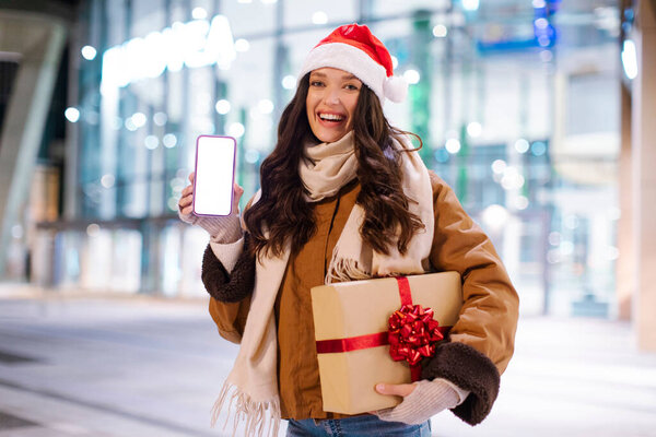 Новогоднее мобильное предложение. Возбужденная леди показывает чистый экран мобильного телефона и держит подарочную коробку, стоя перед торговым центром, макет для рекламы