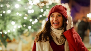 Heyecanlı bayan ışıklarla süslenmiş Noel ağacına karşı duruyor, Noel arifesinde akşam vakti yürüyor, şehrin şenlikli atmosferinin tadını çıkarıyor.