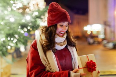 Kış tatili konsepti. Şaşırdım. Noel hediyesini açık hava Xmas kutlamalarında açan ve festival dekorasyonlarına karşı duran bir kadın.
