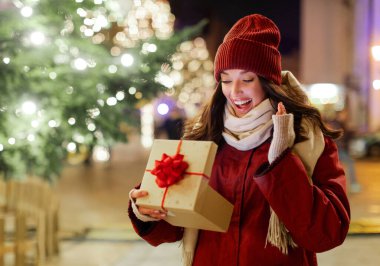 Heyecanlı bayan hediye kutusunu açıyor ve seviniyor, süslenmiş Xmas ağacının önünde duruyor çelenk ışıklarıyla, bedava uzay
