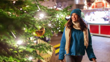 Neşeli bir kadın Noel pazarında yürürken süslenmiş Noel ağacının önünde çelenk ışıklarıyla dikilirken.