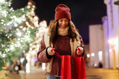 Noel hediyeleriyle dolu bir çantaya bakan mutlu bir kadın Noel pazarında dikiliyor ve köknar ağacıyla süsleniyor.