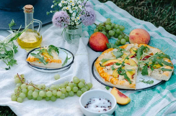Pizza with chicken, peaches, cream cheese and arugula. Picnic on the grass with pizza, grapes, peaches and flowers