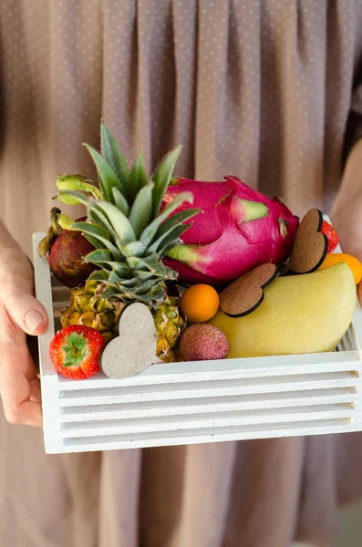 Fruit creative serving. Woman\'s hands holding a wooden box with exotic fruits. Romantic gift. Passion fruit, strawberries, pitahaya, lychee, mango