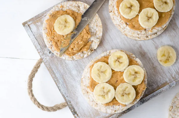 Healthy breakfast with rice cakes with peanut butter and slices of banana on white wooden table. Top view.