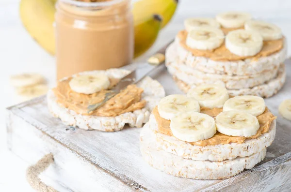 ピーナッツ バターと白の木製テーブルの上にバナナのスライス餅入りの健康的な朝食 — ストック写真