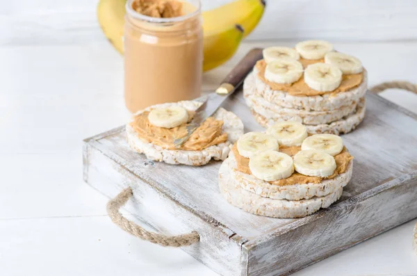 ピーナッツ バターと白の木製テーブルの上にバナナのスライス餅入りの健康的な朝食 — ストック写真