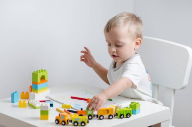 Renkli plastik bir sürü ile oynayan küçük sarışın çocuk, bir oyuncak tren ile engeller ve masada çizer. Küçük çocuk bir sandalye beyaz masada oturur.