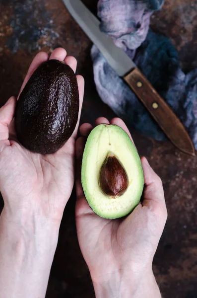 Top down view, woman hands with pink nails holding two whole dark ripe avocados over white boards desk.