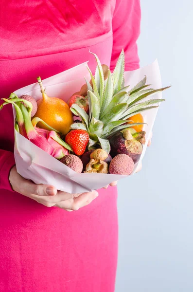 Edible arrangements, edible exotic fruit bouquet. Pineapple, granadilla, litchi, strawberry, pitahaya, mangosteen in the hands of a women in a pink dress.