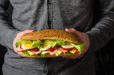 Adam elinde yumurta, peynir, et ve salata ile büyük bir sandviç tutar.