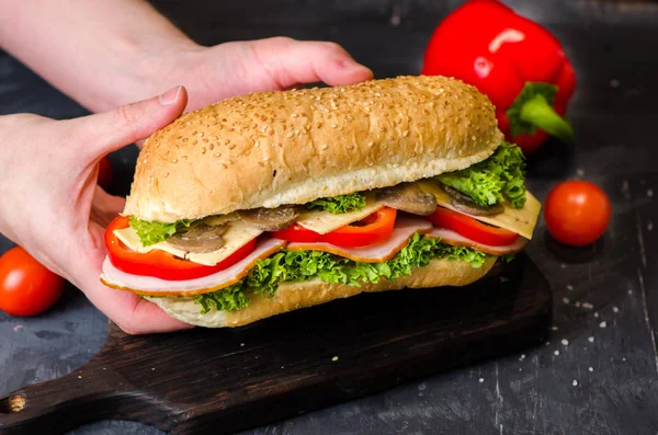 美味的三明治，包括火腿、奶酪、红辣椒、蘑菇和沙拉，摆满新鲜蔬菜的木桌上。手拿三明治 — 图库照片