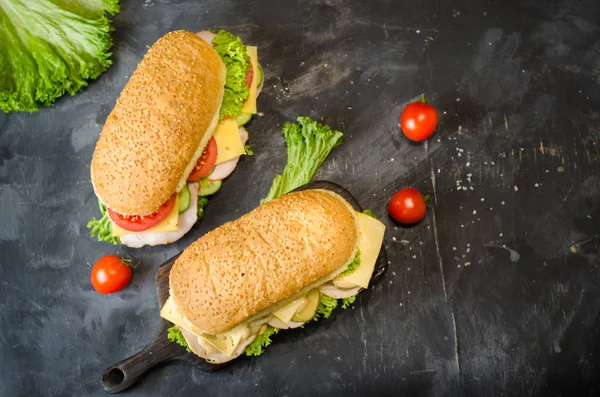 Zwei leckere Sandwiches mit Schinken, Käse, Tomaten, salzigen Gurken und Salat über einem Holzbrett auf schwarzem Hintergrund. Raum für Text. — Stockfoto