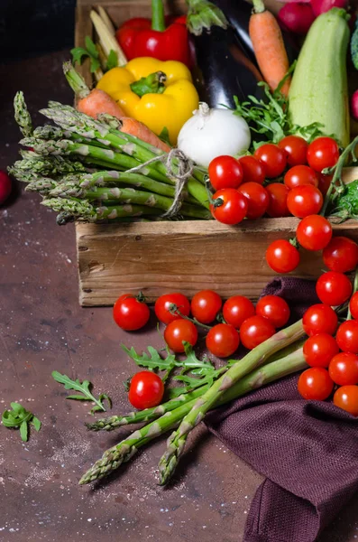 Box with farm vegetables on a dark background. Place for text. Cherry tomatoes, asparagus, broccoli, peppers, eggplants, zucchini, carrots and onions. — Stock Photo, Image
