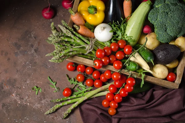 Κουτί με λαχανικά φάρμας σε σκοτεινό φόντο. Θέση για κείμενο. Ντοματίνια, σπαράγγια, μπρόκολο, πιπεριές, μελιτζάνες, κολοκυθάκια, καρότα και κρεμμύδια. — Φωτογραφία Αρχείου