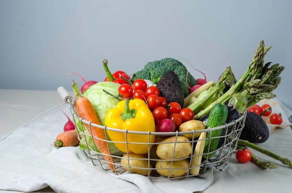 Fresh vegetables in a metal basket on the table. Cherry tomatoes, asparagus, broccoli, potato, peppers, radish, eggplants, zucchini, carrots and onions. — Stock Photo, Image